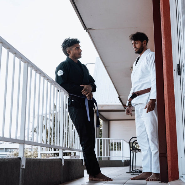 Two martial artists in gis on a balcony; one in black with a blue belt, the other in white with brown belt.